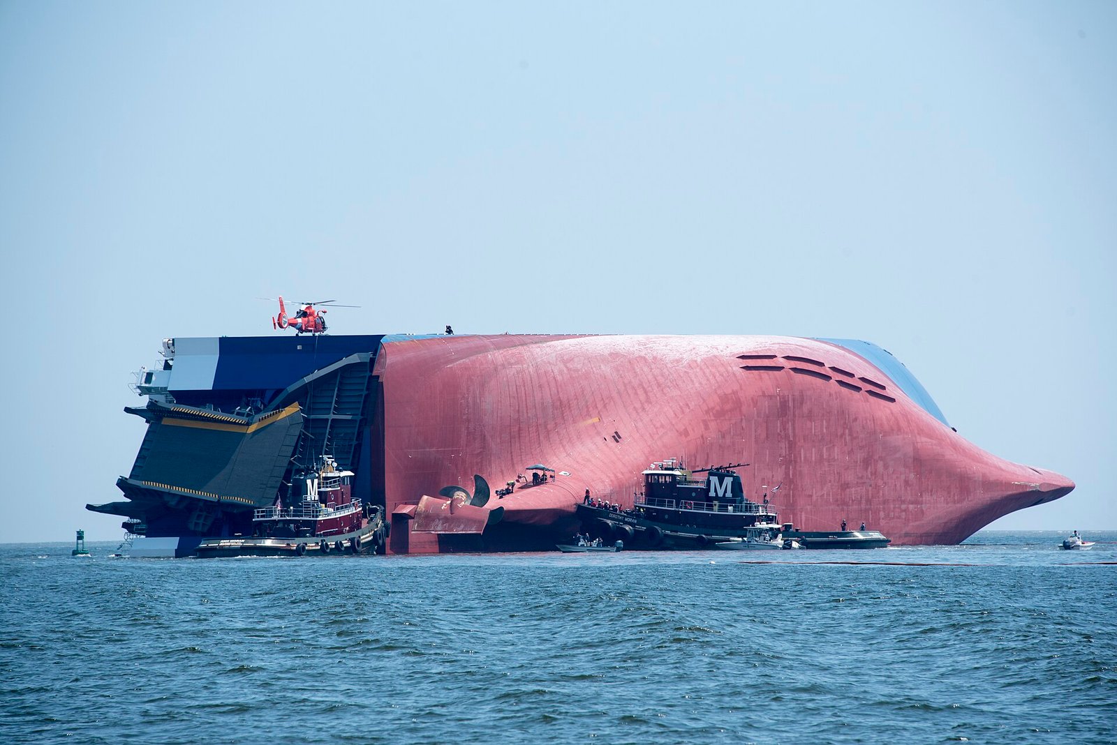 A Coast Guard Air Station Savannah MH-65 Dolphin helicopter rests on the side of the Golden Ray , a 656-foot vehicle carrier, to drop off supplies for Coast Guard crews and port partners who attempt to locate and rescue the remaining four crewmembers aboard the Golden Ray , Sept. 9, 2019, in St. Simons Sound, near Brunswick, Georgia. A Coast Guard Air Station Savannah MH-65 Dolphin helicopter aircrew, Station Brunswick boat crews and other port partners rescued 20 people the morning of Sept. 8, after it was reported the vessel was disabled and listing. The tugs Ann Moran (left) and Dorothy Moran (right) are attending.